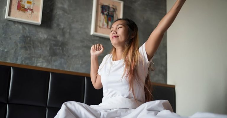 一名女子在早晨醒來,坐在床上、微笑伸展雙臂。