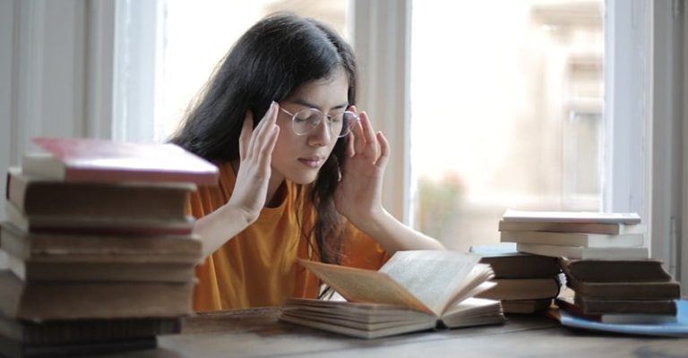 一位年輕女子在堆滿書本的桌子前閱讀，她雙手按壓太陽穴，表現出頭部不適的狀況。