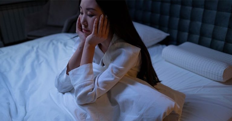 一名失眠的女子坐在床上，表情疲憊，夜晚房間昏暗。