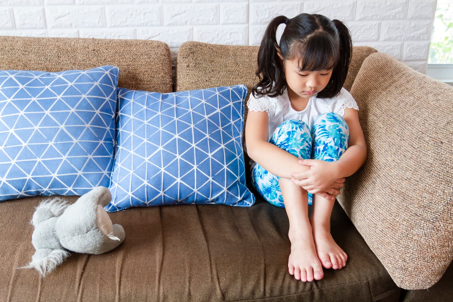 一位年幼的女孩帶著深思和沮喪的表情，孤獨地坐在沙發角落。這個場景凸顯了可能的自閉症症狀，反映出她的孤獨感和內在情緒波動。
