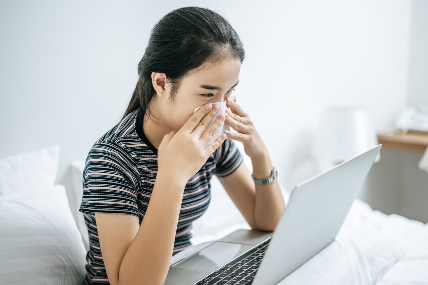 一名年輕女子坐在床上用紙巾擦鼻涕，一邊看著筆記型電腦。
