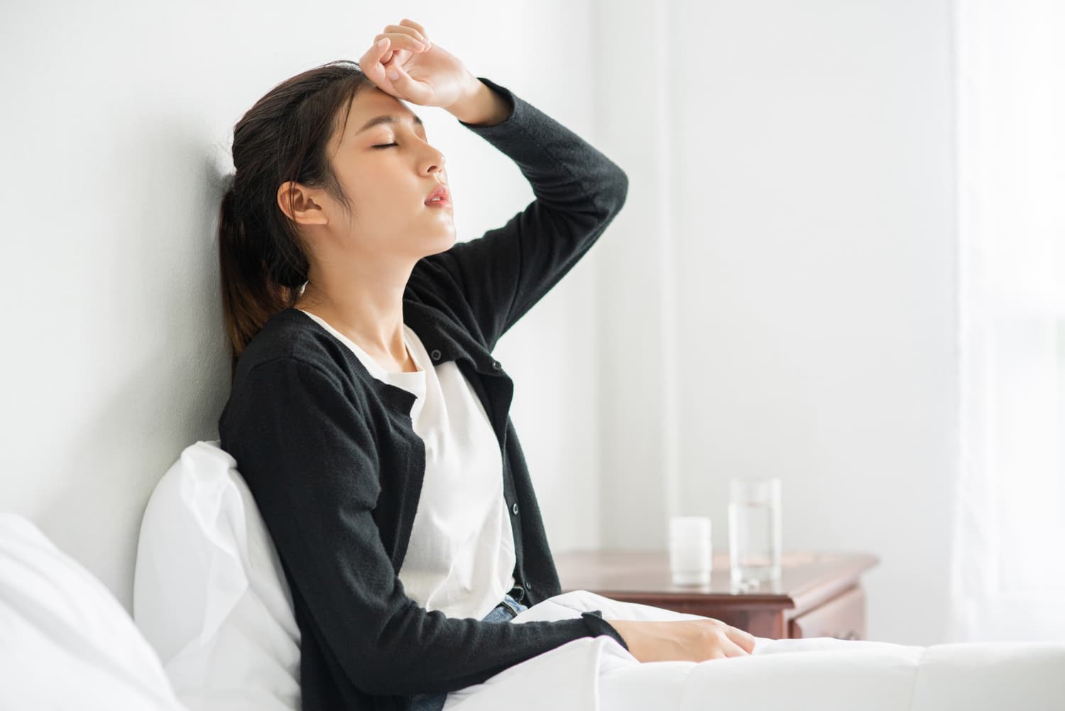 一位穿著黑色外套的女子坐在床上，一隻手放在額頭上，看起來疲憊和虛弱，這可能是血液循環不足或頭痛的症狀。