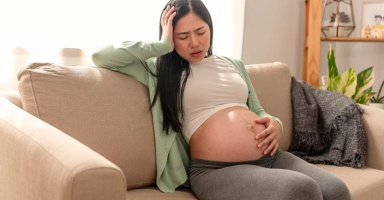 懷孕的女性坐在沙發上，表情痛苦，一手扶著肚子、一手撐著頭，表現出孕期不適或產前憂鬱症的跡象。