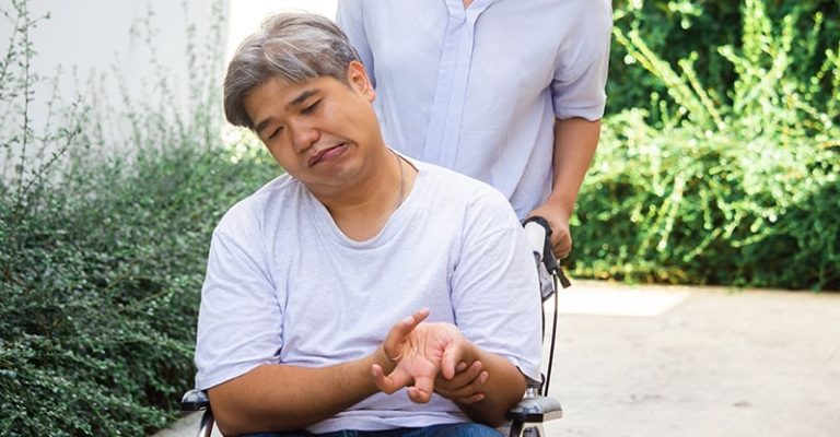 一位坐在輪椅上的中年男子顯得身體無力，可能因中風導致行動不便，左側身體僵硬、臉歪嘴斜。