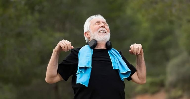 一名年長男子在戶外活動，舉起雙手、脖子掛著毛巾和耳機，露出微笑並享受陽光。