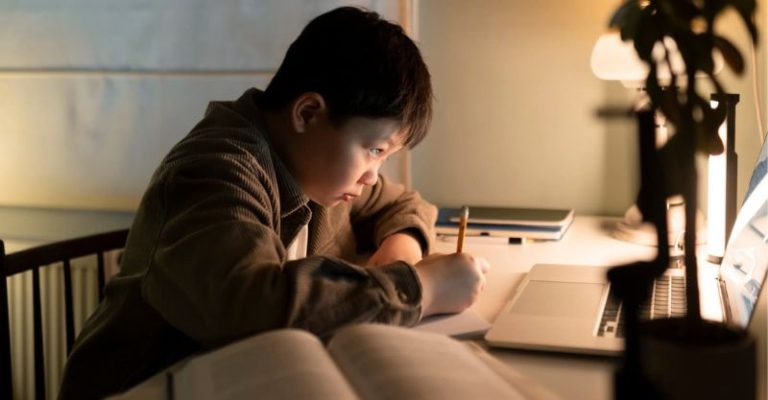 男學生晚上在書桌前使用筆電寫功課，神情專注，桌面擺放書籍與檯燈。