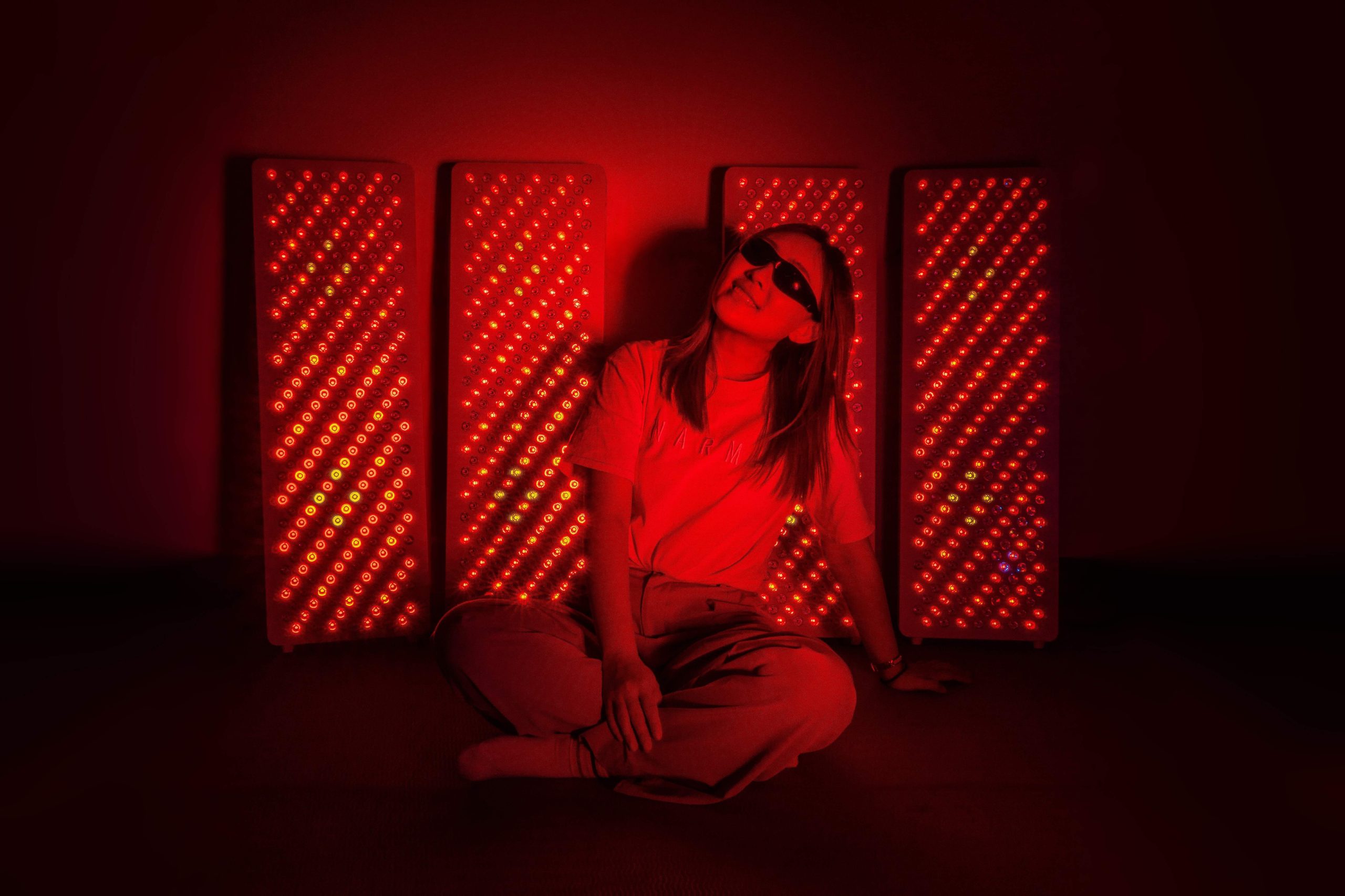 一名女子坐在紅光儀前，穿著輕便衣物並戴著護目鏡進行全身紅光照射，紅光面板排列在背後，用於促進修復、舒緩肌肉與聲帶保養。