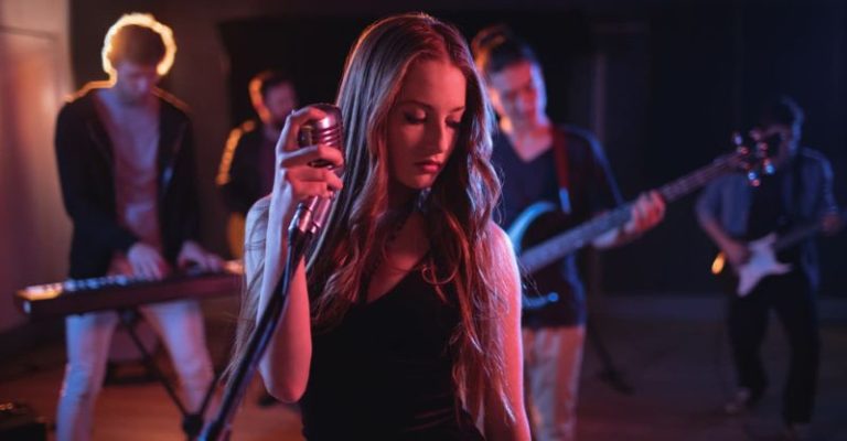 一名女歌手在舞台上手握麥克風,與樂團成員一同表演,背景有鍵盤手、吉他手與貝斯手,燈光營造出現場演唱氛圍。