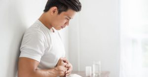 一名年輕男子坐在床邊雙手抱腹，表情痛苦，疑似因脹氣導致腹部不適。