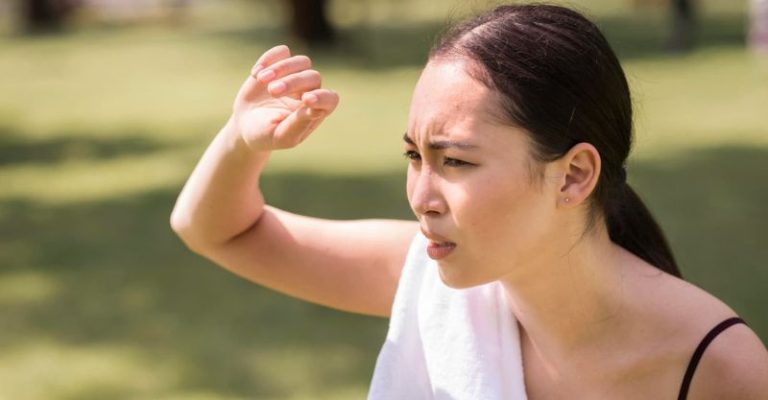 一名女子在烈日下遮陽皺眉,身上掛著毛巾,表情顯露不適,呈現炎炎夏日容易中暑的情境。