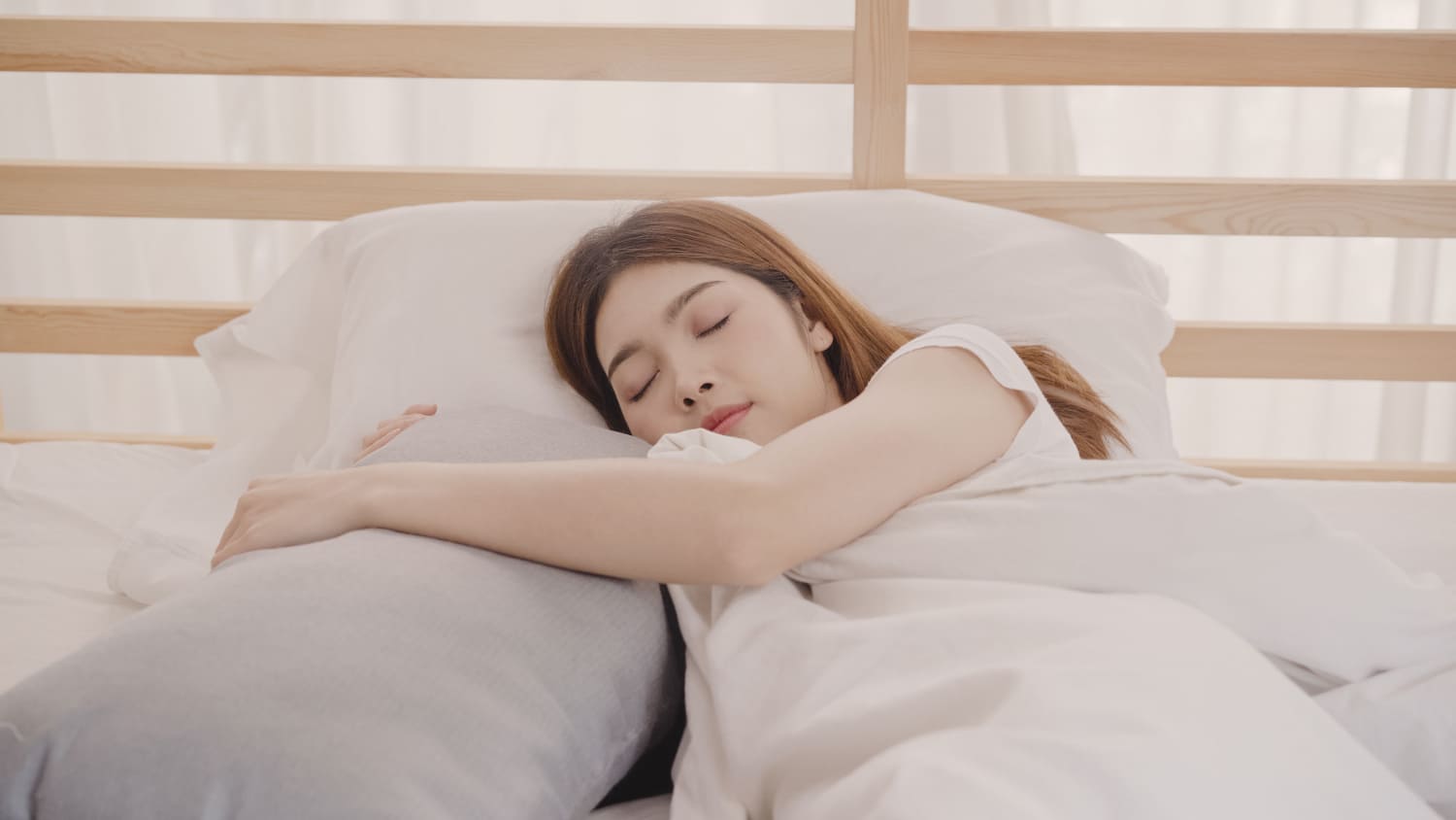 一位女子擁抱長枕安穩入睡,臉上帶著淡淡微笑,陽光透過窗簾灑進室內,營造出溫暖柔和的早晨氛圍。