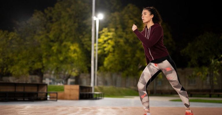一位女子身穿運動服與亮色運動鞋,在夜晚的戶外跑道上進行暖身運動,背景為燈光明亮的公園與樹木,展現夜間鍛鍊的專注與活力。